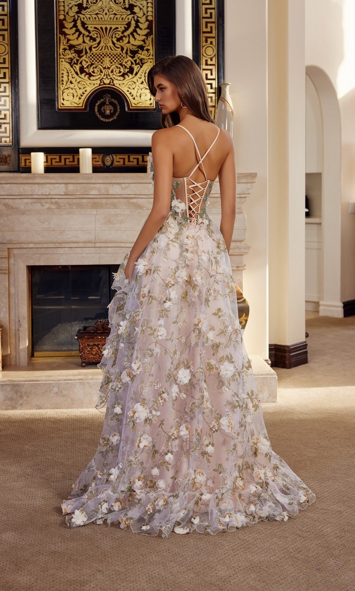 A woman stands facing away in a carpeted room with a fireplace, wearing the Nox Anabel G1783 long prom dress featuring 3D floral appliques and a lace-up back.